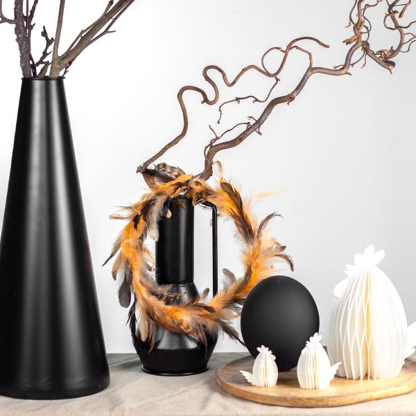 Dekorative Szene mit schwarzen Vasen, Federkranz, Osterei und Papierhühnern auf einem Holzteller.