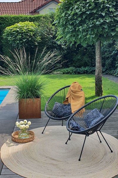 Modernes Gartenmöbel Set mit Designerstühlen aus Metall, rundem Teppich und hohem Pflanzkübel auf einer Holzterrasse neben Rasen und Pool.