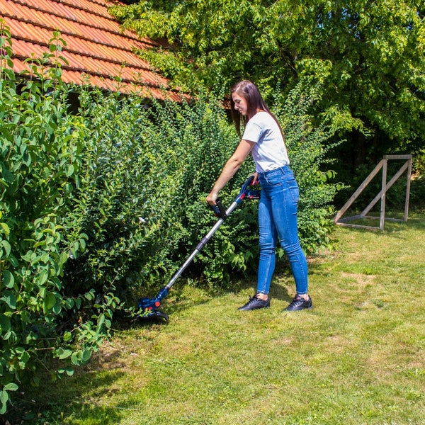Eine Frau benutzt einen Akku-Rasentrimmer zum Schneiden von Gras und Rasenkanten an einer grünen Hecke im Garten.