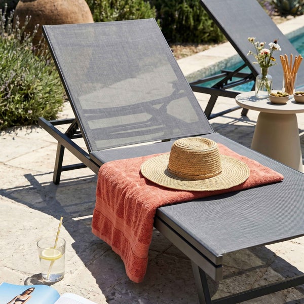 Moderne Gartenliege aus Metall und Textilgewebe auf einer hellen Terrasse am Pool mit Handtuch und Strohhut.