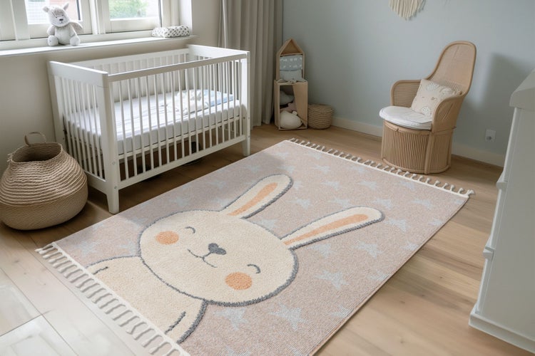 Kinderzimmer mit weißem Gitterbett, Rattan-Sessel und rechteckigem Teppich mit Hasenmotiv und Sternenmuster auf hellem Holzboden.