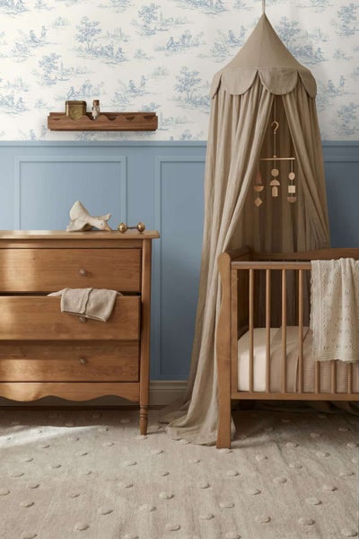 Ein gemütliches Babyzimmer mit Holzkommode, Gitterbett mit Betthimmel, strukturiertem Teppich und einer Kombination aus Tapete und Wandverkleidung.
