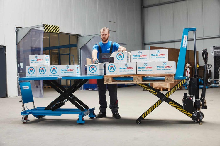Mitarbeiter belädt einen Scherenhubwagen und einen Scherenhubtischwagen mit HanseLifter Kartons in einer hellen Lagerhalle.