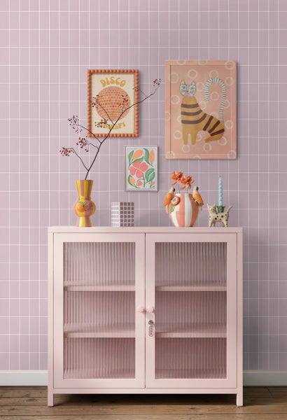 Rosa Metallschrank mit Riffelglastüren und Regalen vor einer Gittertapete. Dekovasen, Kerzenständer und Bilderrahmen ergänzen das moderne Design.