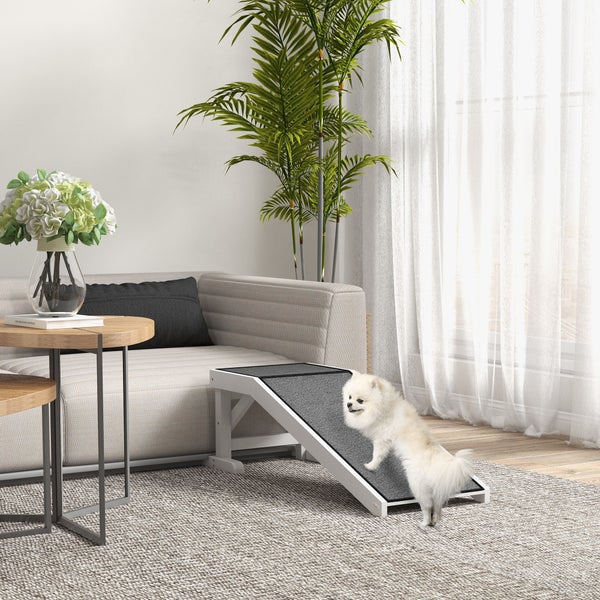 Ein kleiner weißer Hund nutzt eine weiße Hunderampe aus Holz mit grauer Antirutschfläche in einem hellen Wohnzimmer neben einem Sofa.