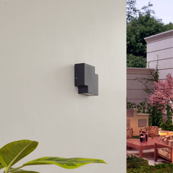 Moderne schwarze Außenwandleuchte in geometrischem Design an einer hellen Hauswand im Gartenbereich.