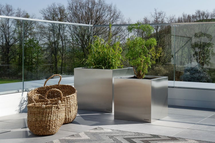 Zwei quadratische Pflanzkübel aus Edelstahl mit grünen Pflanzen und drei geflochtene Weidenkörbe auf einer modernen Terrasse mit Glasgeländer.