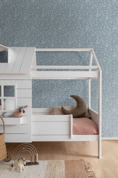Helles Kinderzimmer mit einem weißen Hausbett aus Holz vor einer blauen Blütentapete und hellem Parkettboden.