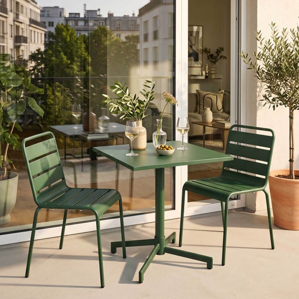 Moderner, heller Balkon mit zwei grünen Metallstühlen und einem passenden quadratischen Tisch. Dekoriert mit Pflanzen, Weingläsern und Oliven.