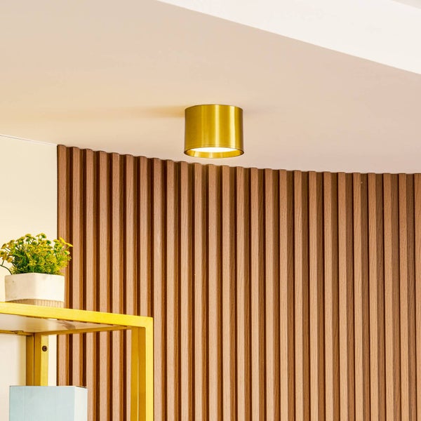 Zylindrischer Deckenstrahler in Goldoptik montiert an der Decke vor einer Wand mit Holzlamellen.