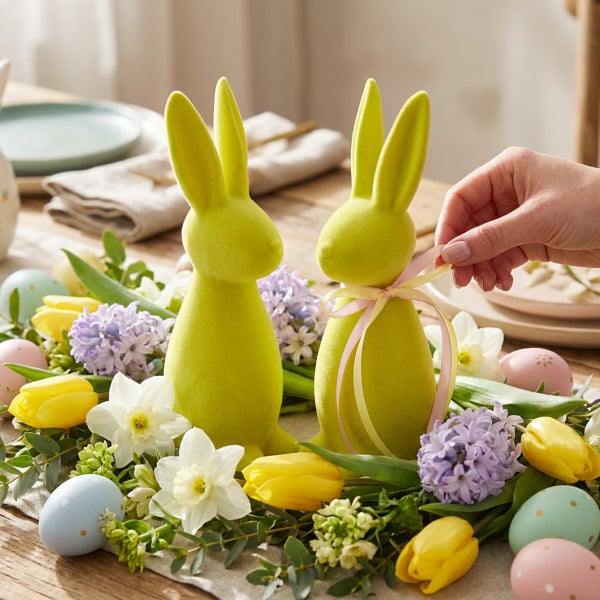 Zwei dekorative Osterhasen aus beflocktem Material mit einer Schleife auf einem frühlingshaften Tischgesteck mit Blumen und Ostereiern