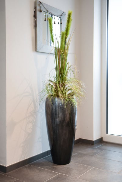 Hohe, dunkelgraue Boden-Vase mit künstlichem Ziergras in einem hellen, modern gestalteten Raum auf dunklen Bodenfliesen.