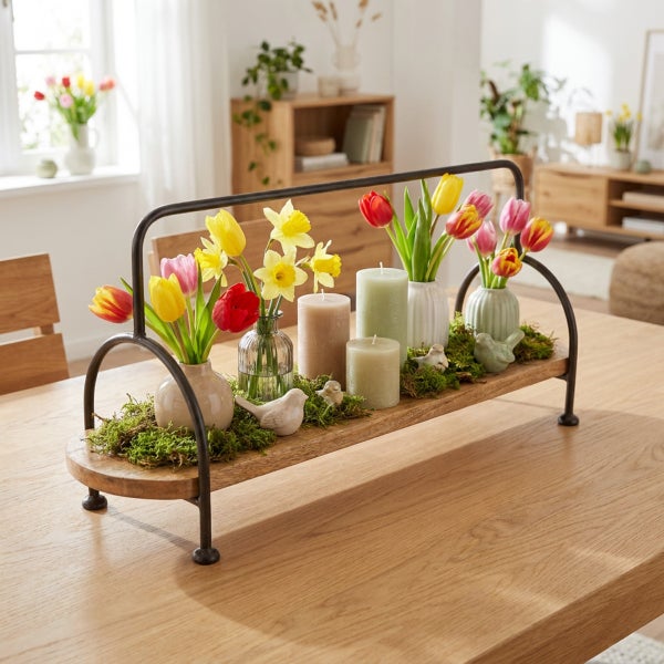 Frühlingshafte Tischdekoration mit Holztablett, Metallgriff, Vasen mit Tulpen und Narzissen, Stumpenkerzen sowie kleinen Deko Vögeln auf Moos.