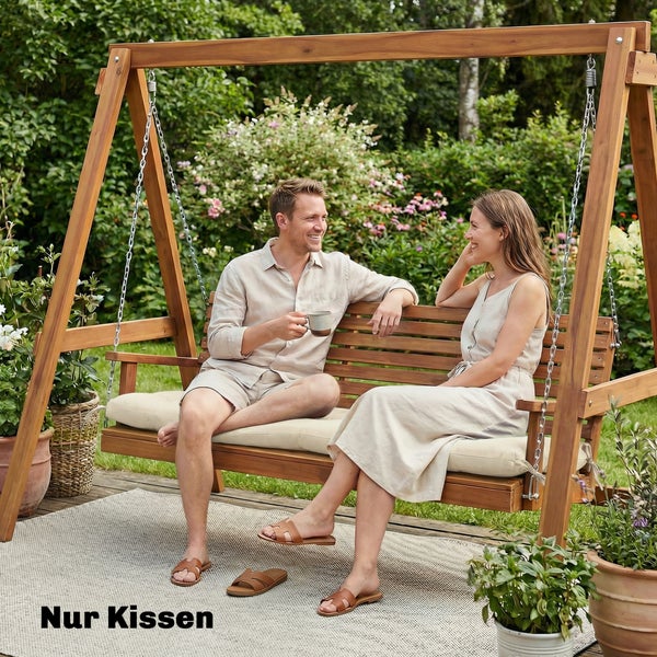 Paar sitzt auf einer hölzernen Hollywoodschaukel mit beigen Sitzkissen in einem hellen Garten auf einem Outdoorteppich.