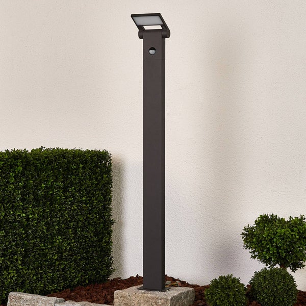 Moderne Außenstandleuchte mit Bewegungsmelder auf einem Steinsockel im Garten neben einer Hecke.