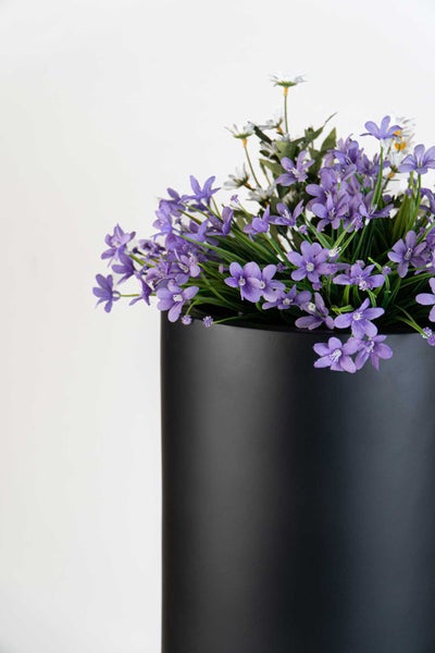 Künstliche Blumen in einem schwarzen zylindrischen Pflanzgefäß mit matter Oberfläche.