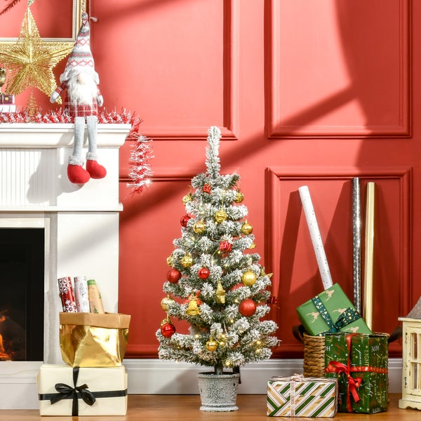 Künstlicher beschneiter Weihnachtsbaum mit roten und goldenen Kugeln und Lichtern neben einem weißen Kamin und verpackten Geschenken.