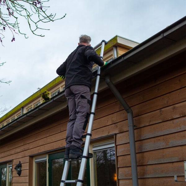 Eine Person steht auf einer Aluminium-Teleskopleiter, um eine Dachrinne an einem Holzhaus mit waagerechter Verschalung zu reinigen.