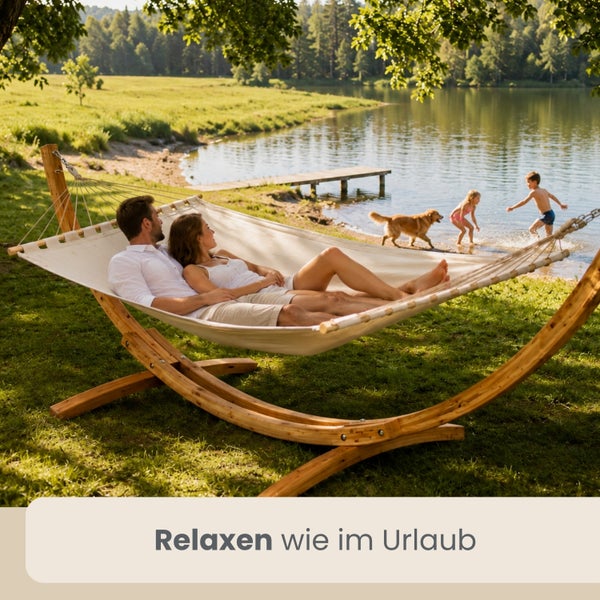 Ein Paar liegt in einer großen weißen Hängematte mit einem Gestell aus Holz auf einer Wiese an einem See.
