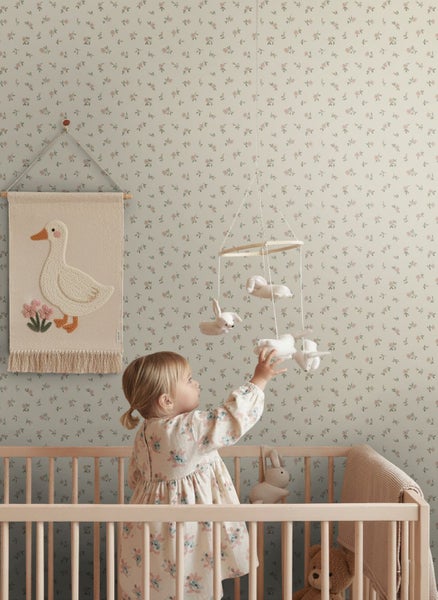 Helles Kinderzimmer mit Holzgitterbett vor Tapete mit floralem Muster, Textilwandbehang mit Ente und Mobile mit Stofftieren.