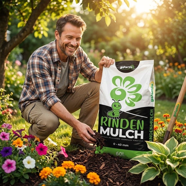 Ein Mann verteilt Galamio Rindenmulch Körnung null bis vierzig Millimeter aus einem sechzig Liter Sack in einem sonnigen Gartenbeet mit Blumen.