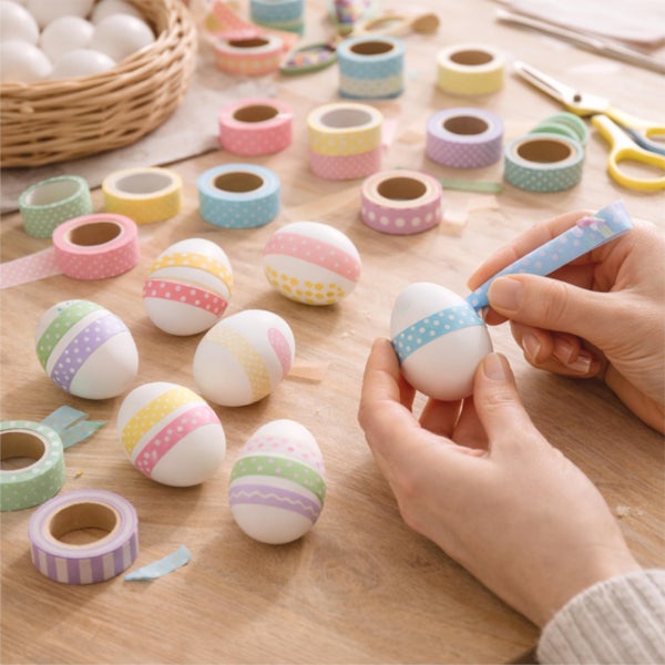 Hände dekorieren weiße Ostereier mit bunt gemustertem Washi-Tape auf einem Holztisch, umgeben von weiteren verzierten Eiern und Kleberollen.