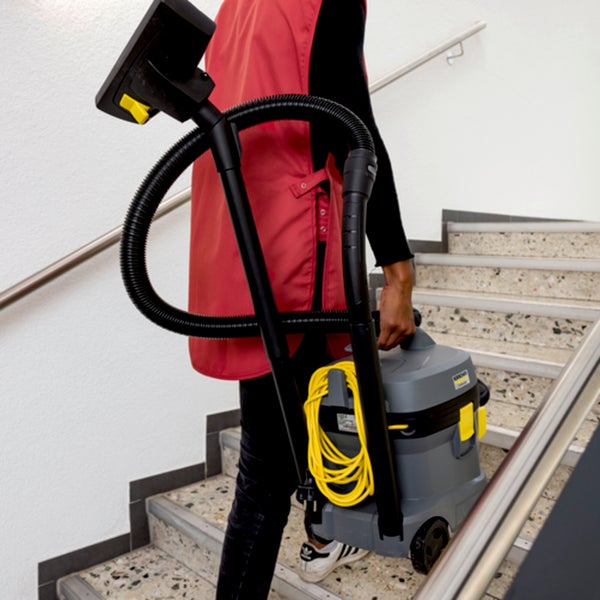 Eine Person trägt einen grauen Trockensauger mit gelbem Kabel an einem Griff eine Treppe hinauf.