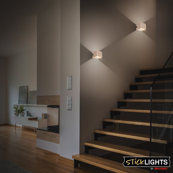 Zwei würfelförmige LED-Wandleuchten an einer Wand neben einer Holztreppe, die ein kreuzförmiges Lichtbild in einem modernen Raum erzeugen.