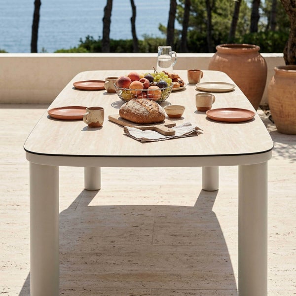 Moderner rechteckiger Gartentisch mit abgerundeten Ecken und dicken zylindrischen Beinen, gedeckt für ein Frühstück auf einer Terrasse mit Meerblick.