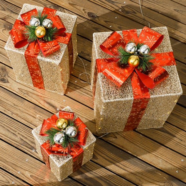 Drei beleuchtete Weihnachtsgeschenke in Gold mit roten Schleifen, Tannenzweigen und Weihnachtskugeln auf einem Holzboden.