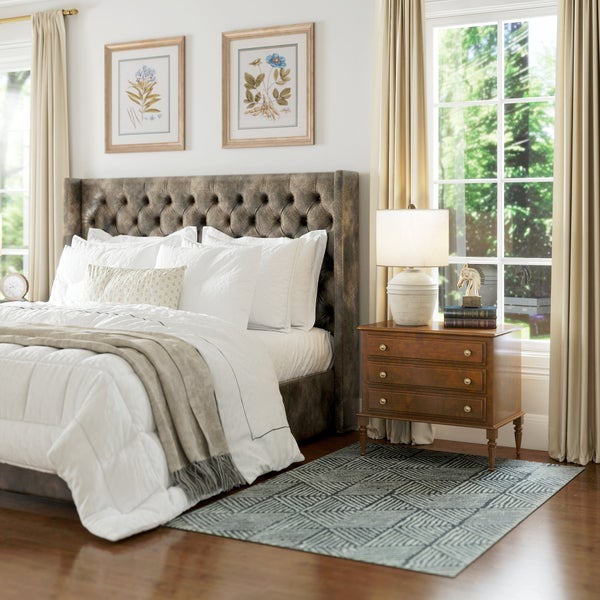 Helles Schlafzimmer mit braunem Polsterbett, weißer Bettwäsche, hölzernem Nachttisch, Tischleuchte, gemustertem Teppich und botanischen Bildern.