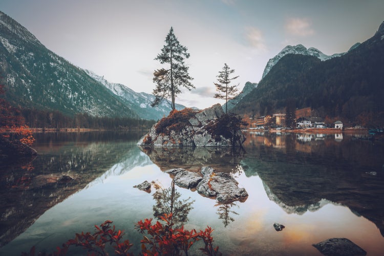 Bergsee mit einer felsigen Insel und zwei Bäumen vor einer alpinen Kulisse bei ruhiger Stimmung.