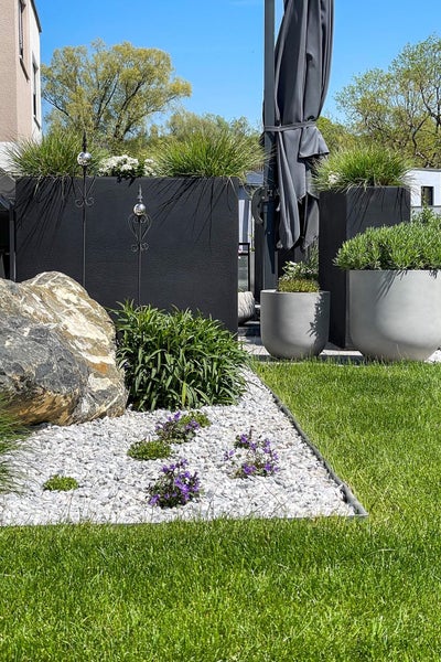 Moderne Gartengestaltung mit Rasenfläche, Beet mit weißem Kies und Naturstein, schwarzen und grauen Pflanzkübeln sowie Sonnenschirm.