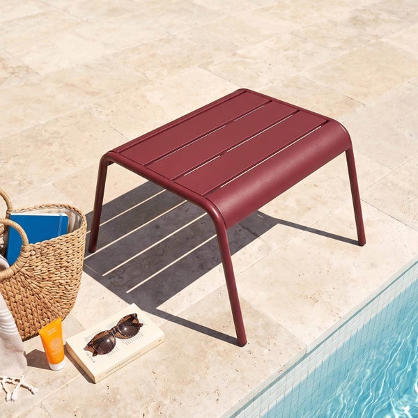 Moderner dunkelroter Beistelltisch aus Metall mit Lamellen auf einer hellen Steinterrasse neben einem Pool im Sonnenlicht.