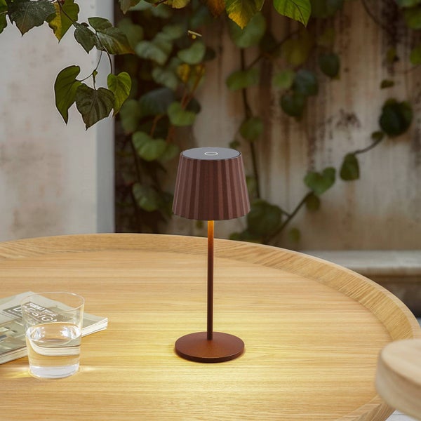 Moderne kabellose LED-Tischleuchte in Rostbraun mit plissiertem Schirm auf einem Holztisch im Außenbereich neben einem Wasserglas.