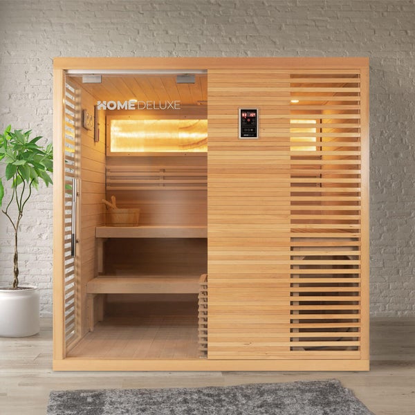 Home Deluxe Infrarotsauna aus Holz mit Glastür und Innenausstattung inklusive Saunaeimer und Kelle vor einer Backsteinwand.
