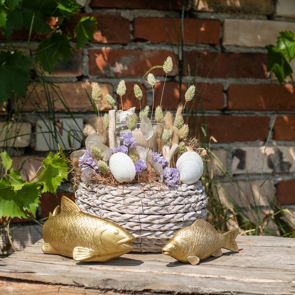 Dekorative Anordnung mit Trockenblumen, Muscheln und goldenen Fischfiguren in einem Weidenkorb