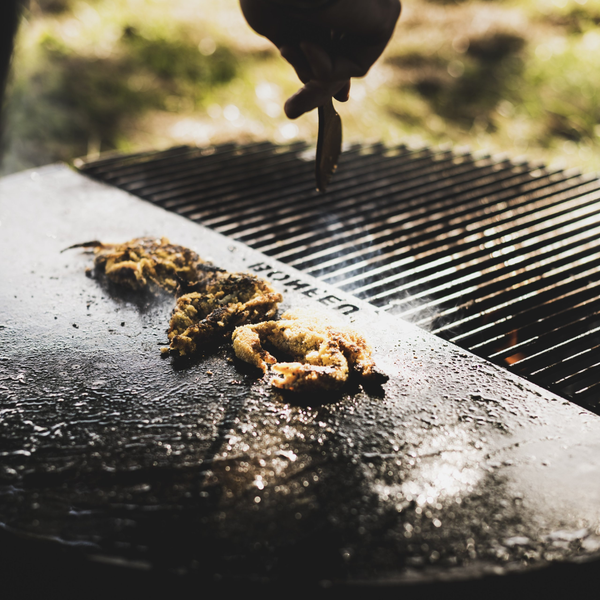 Zubereitung von Speisen auf einer Grillplatte und einem Grillrost aus Metall mit ROHEEN Logo im hellen Außenbereich.