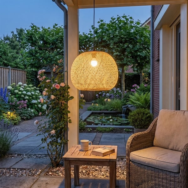 Geflochtene Bambus-Pendelleuchte über Holztisch und Rattansessel auf einer Terrasse, im Hintergrund ein Garten mit Teich bei Dämmerung.