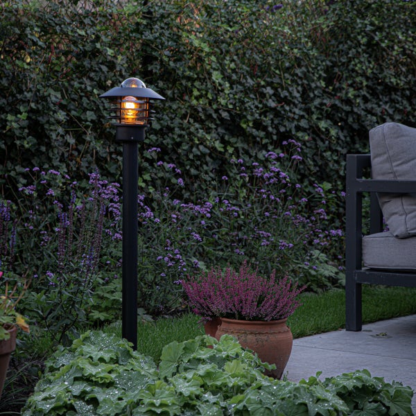 Schwarze Außenstandleuchte in einem bepflanzten Garten neben einer Terrasse mit Gartenmöbeln.