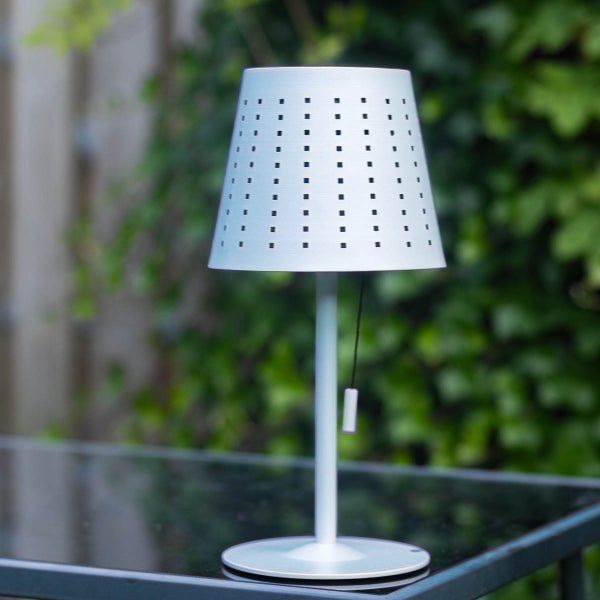 Moderne Außen-Tischleuchte mit perforiertem Lampenschirm und Zugschalter auf einem Tisch im Außenbereich.