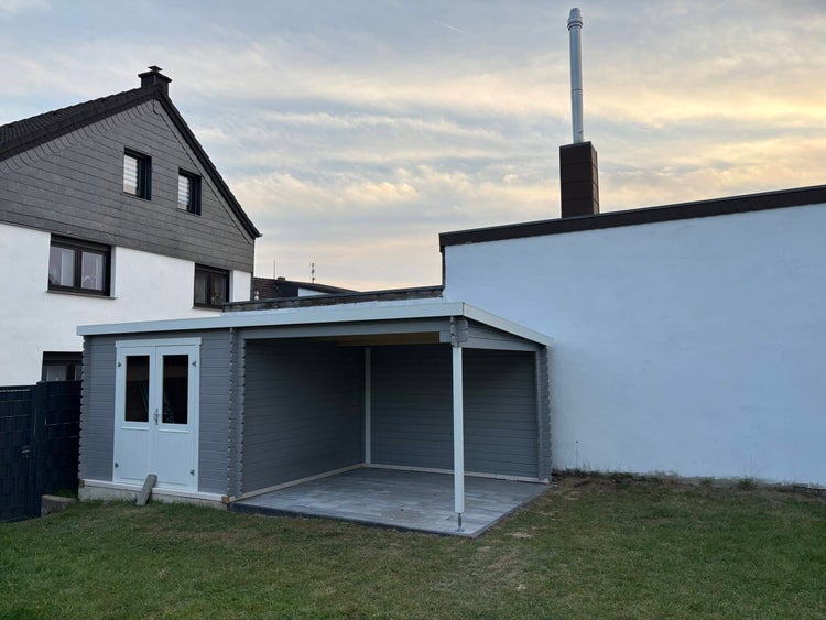 Graues Gartenhaus aus Holz mit weißen Doppeltüren und einem angebauten offenen Unterstand auf einer Rasenfläche.