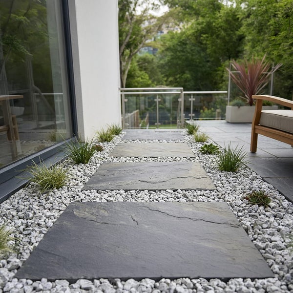 Gartenweg mit Trittplatten aus Schiefer eingebettet in hellem Zierkies entlang einer modernen Hausfassade mit Terrasse und Pflanzen.
