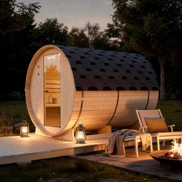 Fasssauna von Home Deluxe aus Holz mit Glasfront auf einer Holzterrasse im Garten, beleuchtet mit Laternen und Feuerstelle am Abend.