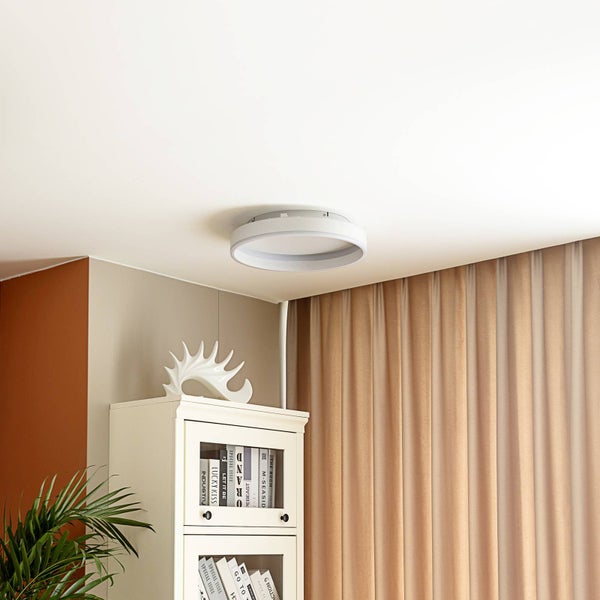Moderne, runde LED-Deckenleuchte in einem hellen Wohnzimmer mit weißem Schrank und beigen Vorhängen.