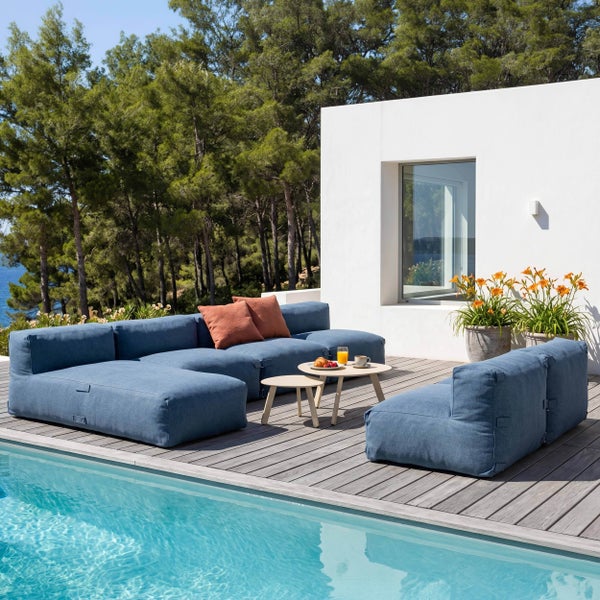 Moderne Outdoor-Lounge am Pool mit blauem Modulsofa, Holzterrasse und runden Couchtischen vor einer bewaldeten Kulisse.