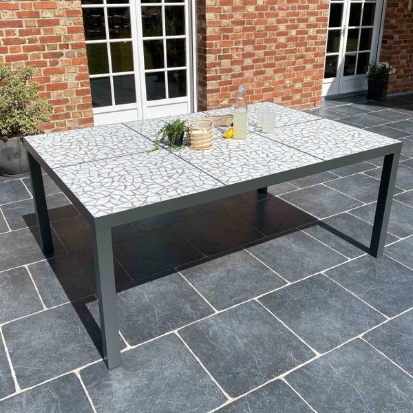 Rechteckiger Gartentisch mit einer hellen Mosaik-Steinplatte und einem anthrazitfarbenen Metallgestell auf einer gefliesten Terrasse.