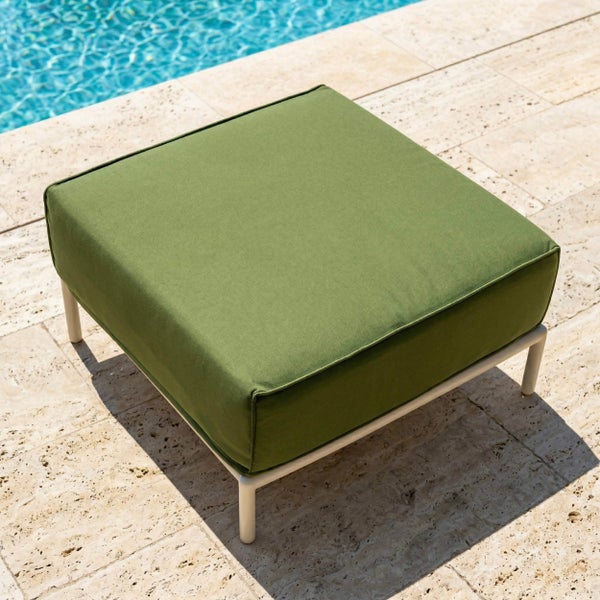 Quadratischer Gartenhocker mit grünem Polster und hellem Metallrahmen auf einer hellen Steinterrasse neben einem Pool.