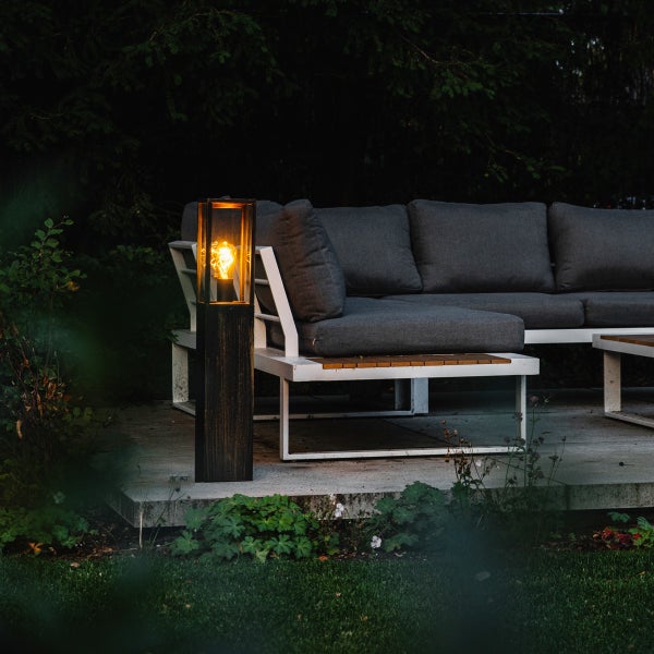 Moderne Außenstehleuchte aus Metall mit Leuchtmittel neben einer Lounge-Sitzgruppe aus Holz und Metall auf einer Terrasse.