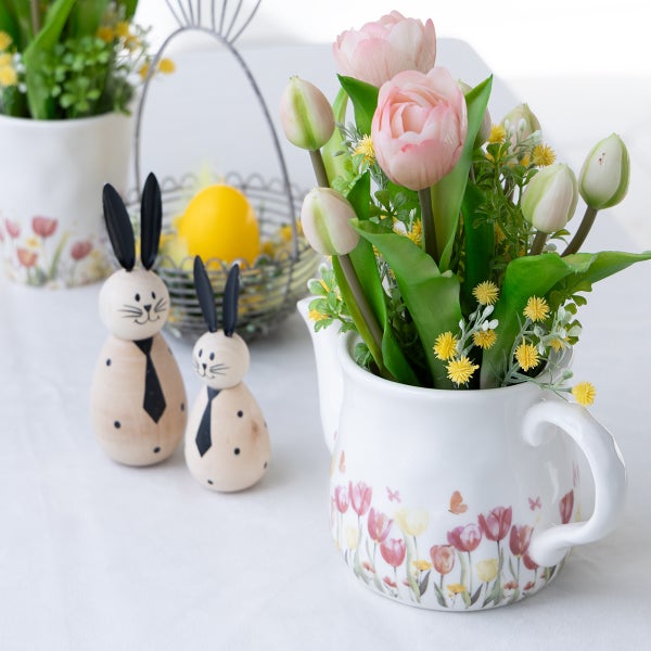 Osterdekoration mit weißem Keramikkrug mit Blumenmuster, künstlichen Tulpen, Holzhasen und Drahtkorb mit gelbem Ei in heller Atmosphäre.